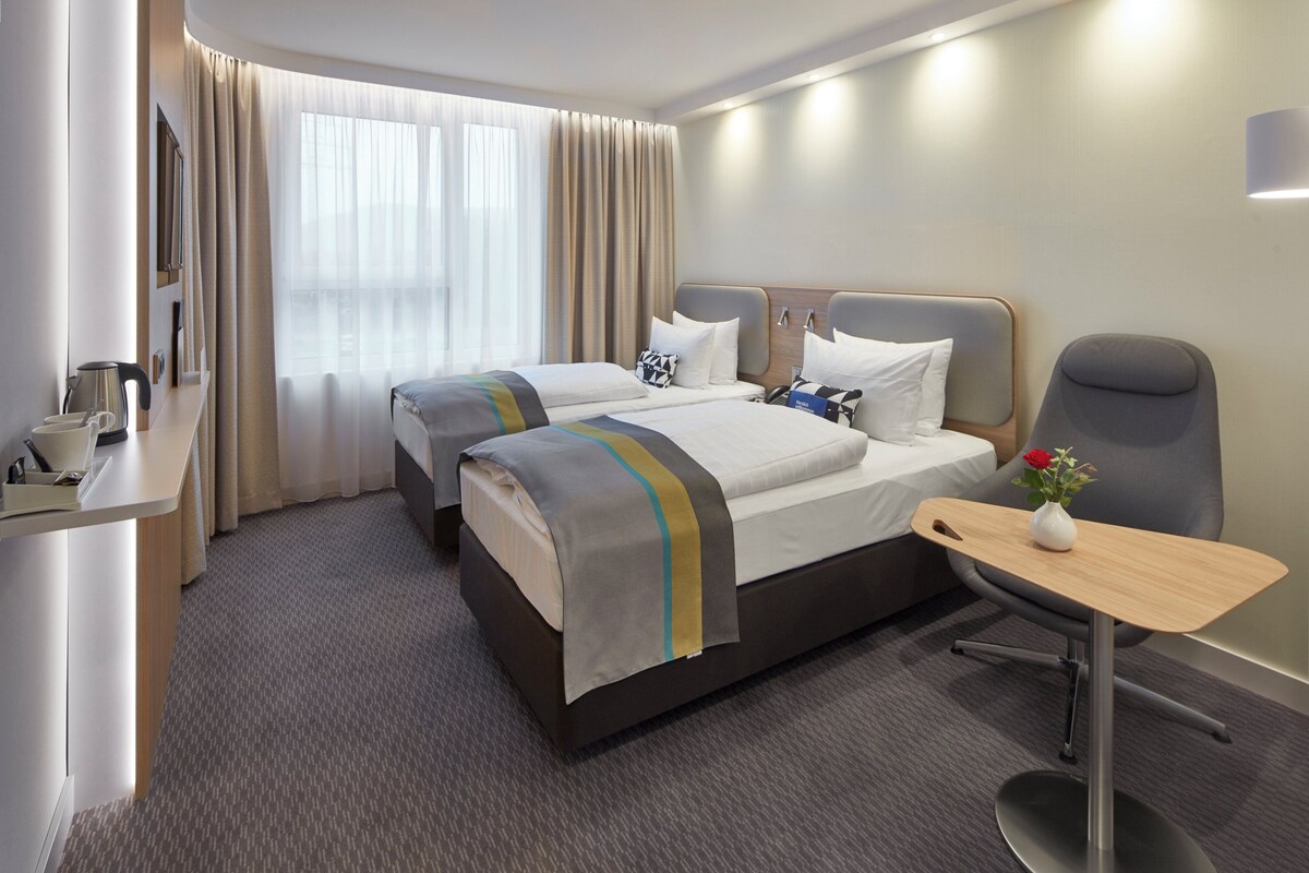 A hotel room with two beds, a desk, a chair, a small table with a vase, and a window with curtains.