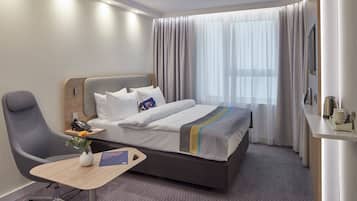 Una habitación de hotel con una cama, un escritorio, una silla, un florero con flores y una ventana con cortinas.