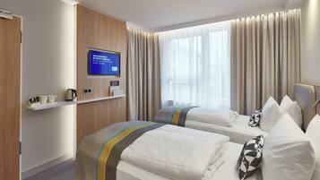 Ein Hotelzimmer mit zwei Betten, einem Fernseher, einem Fenster mit Vorhängen und einem Regal mit einer Kaffeemaschine.