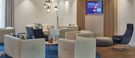 Un moderno vestíbulo de hotel con zona de estar que incluye sofás y sillones, una mesa de centro redonda y un televisor de pantalla plana montado en la pared.