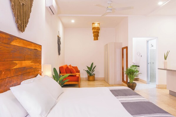 Un dormitorio con cabecera de madera, cama con ropa de cama blanca, un sofá pequeño naranja, una lámpara colgante y una planta en maceta.