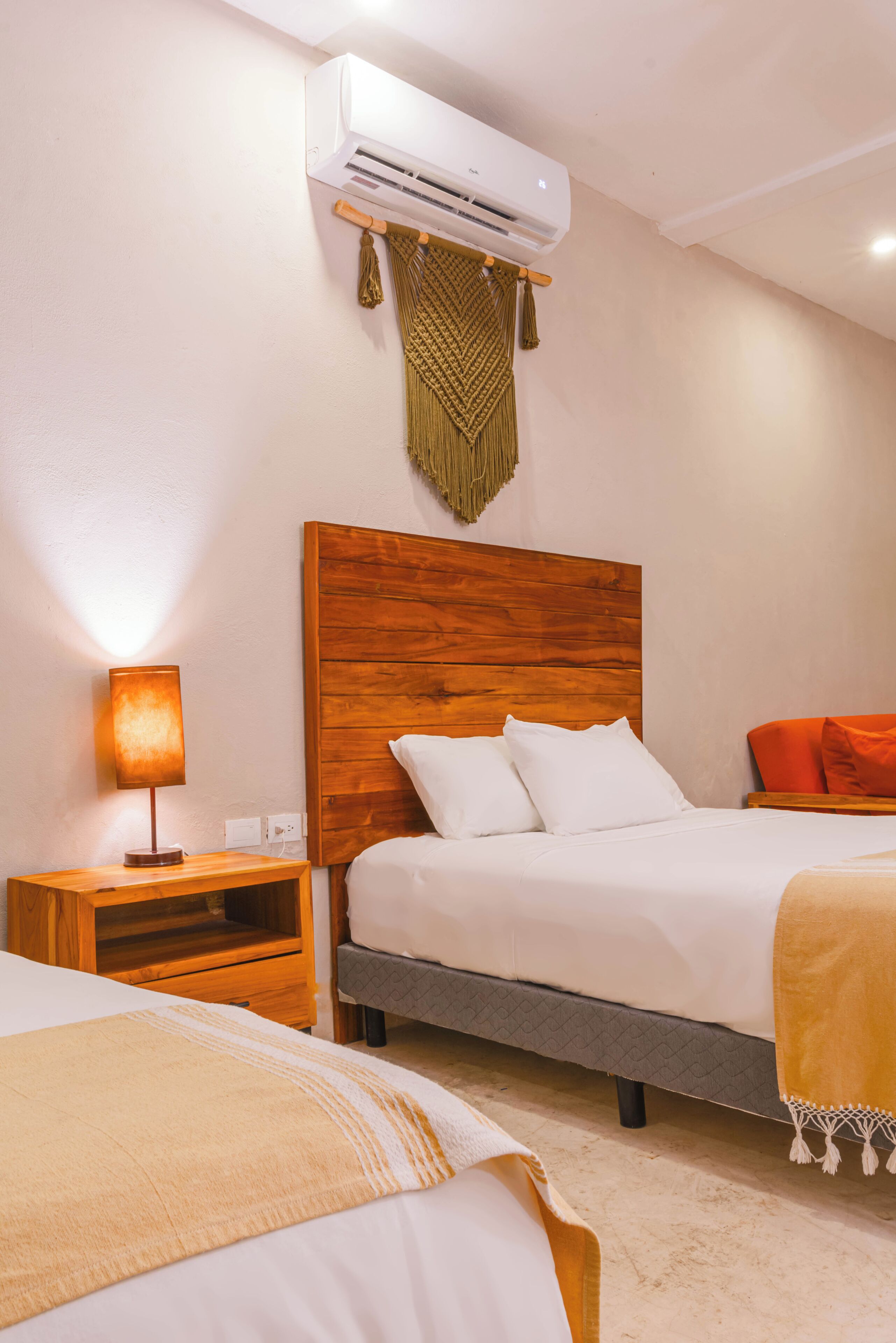 Quarto de hotel com cabeceira de madeira, cama com roupa de cama branca e laranja, mesa de cabeceira com abajur e unidade de ar condicionado.