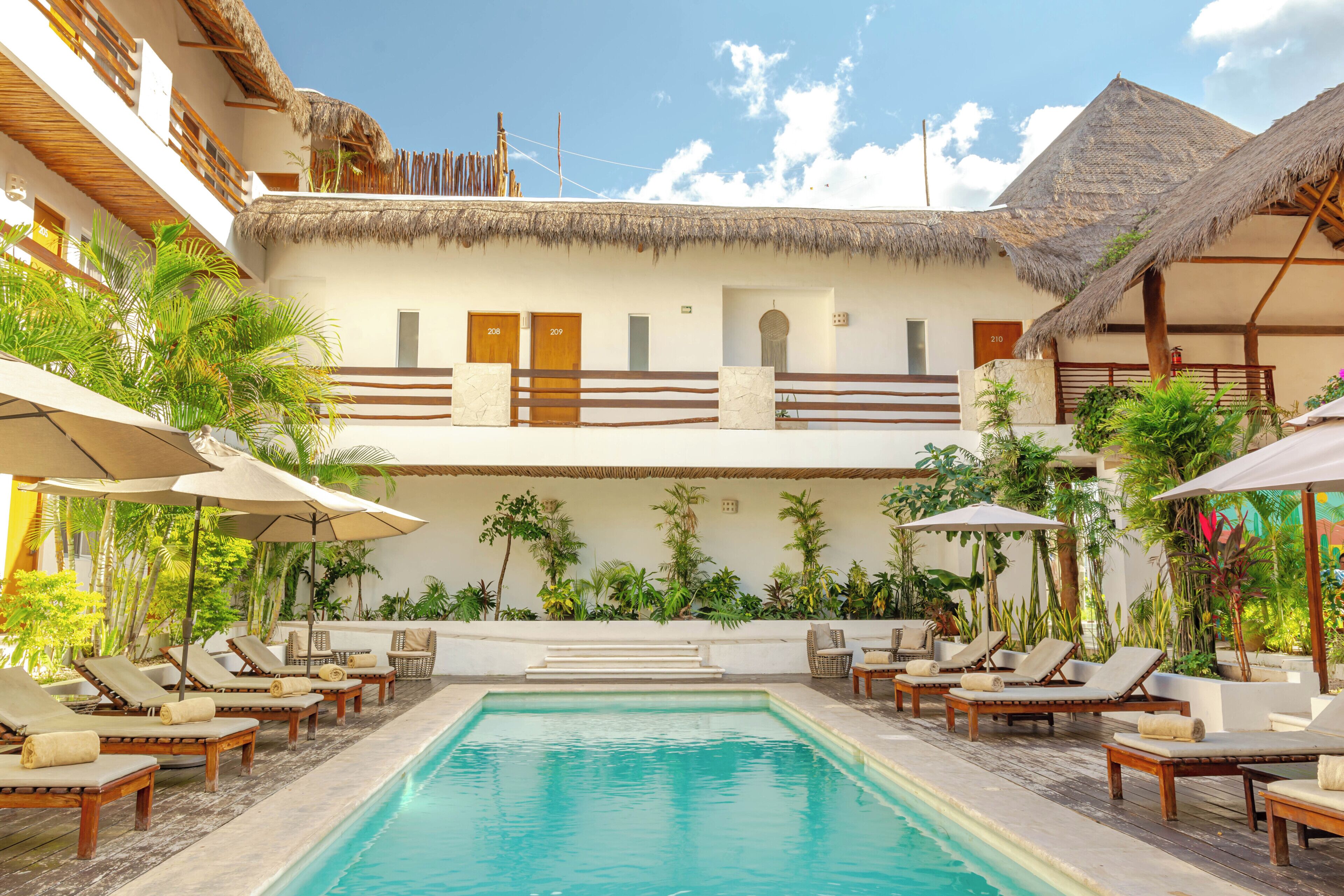 Zona de piscina con sillones de playa y sombrillas, rodeada de exuberante vegetación y un edificio con techo de paja.