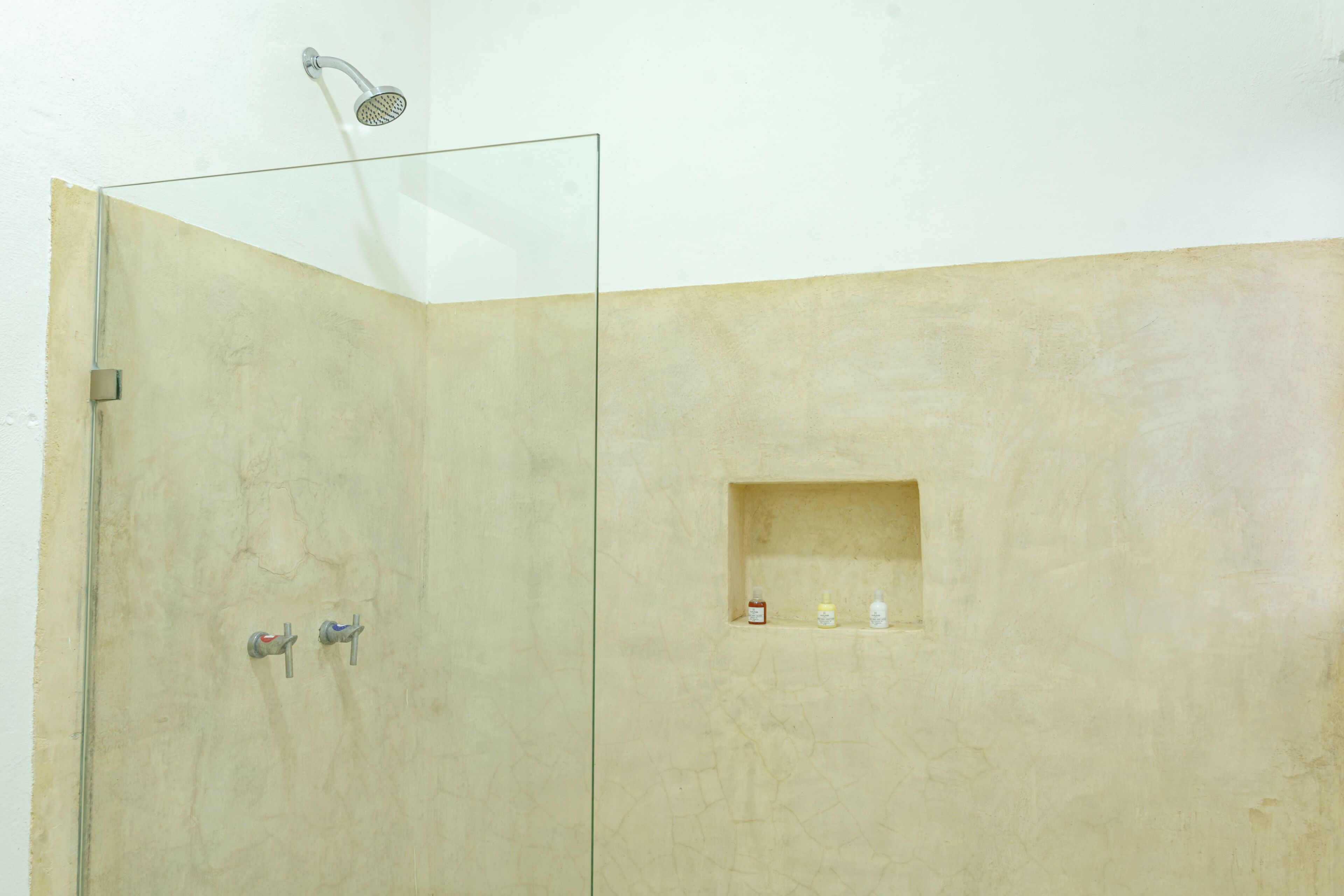 A glass-enclosed shower with a handheld showerhead, a wall-mounted faucet, and a shelf with toiletries.