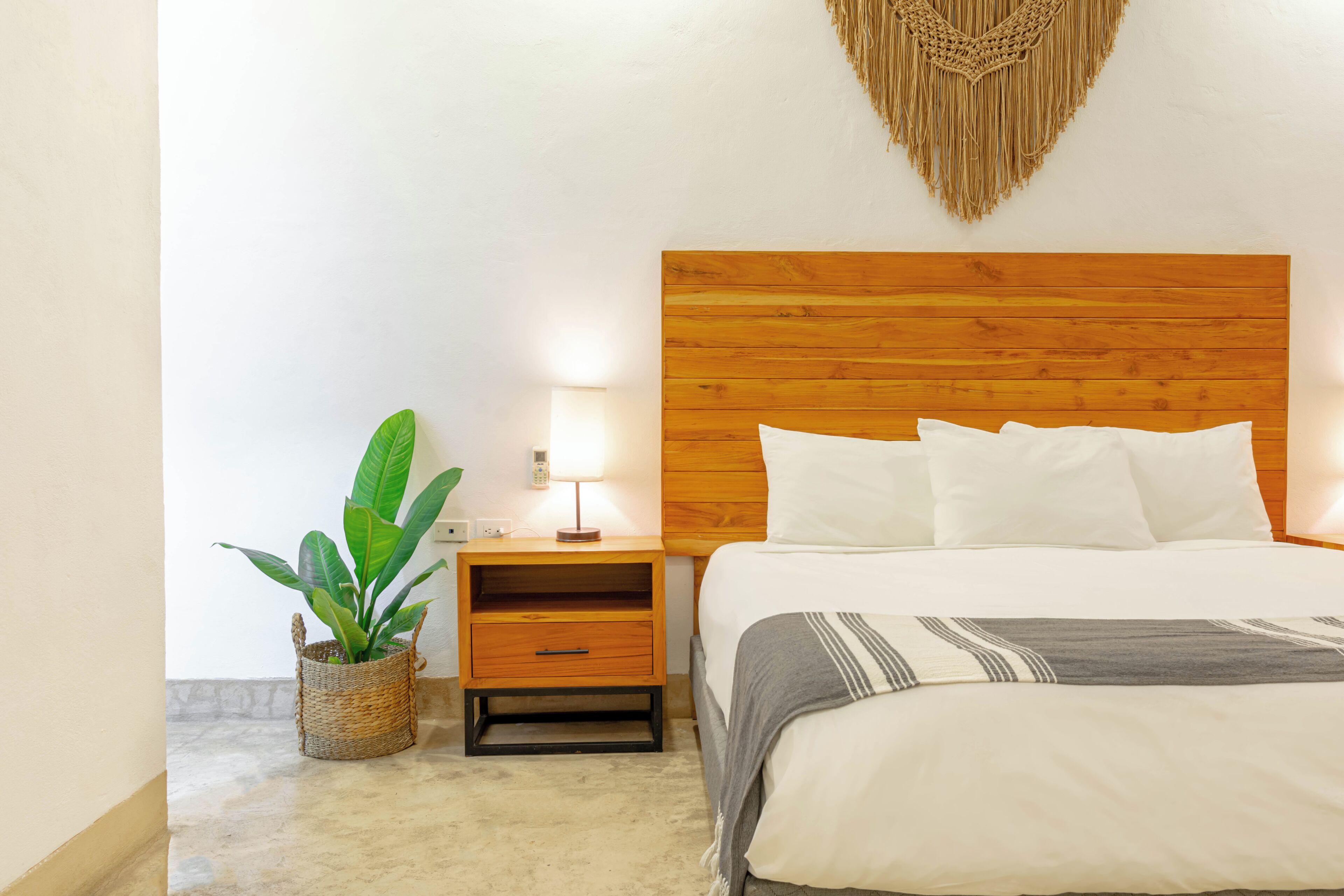Una habitación de hotel con cabecera de madera, cama con ropa blanca, lámpara de noche, una planta pequeña y una cesta tejida.
