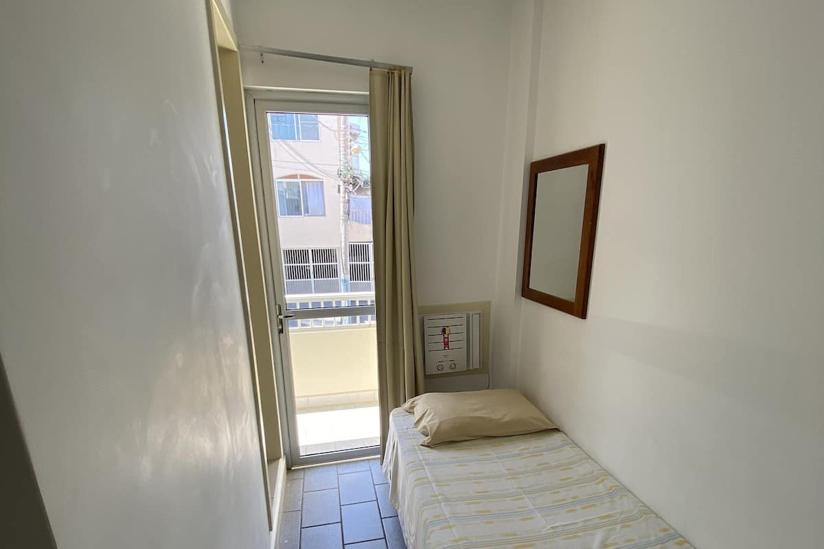 Habitación de hotel estrecha con una cama, una mesita de noche pequeña, un espejo montado en la pared y una ventana con cortina.