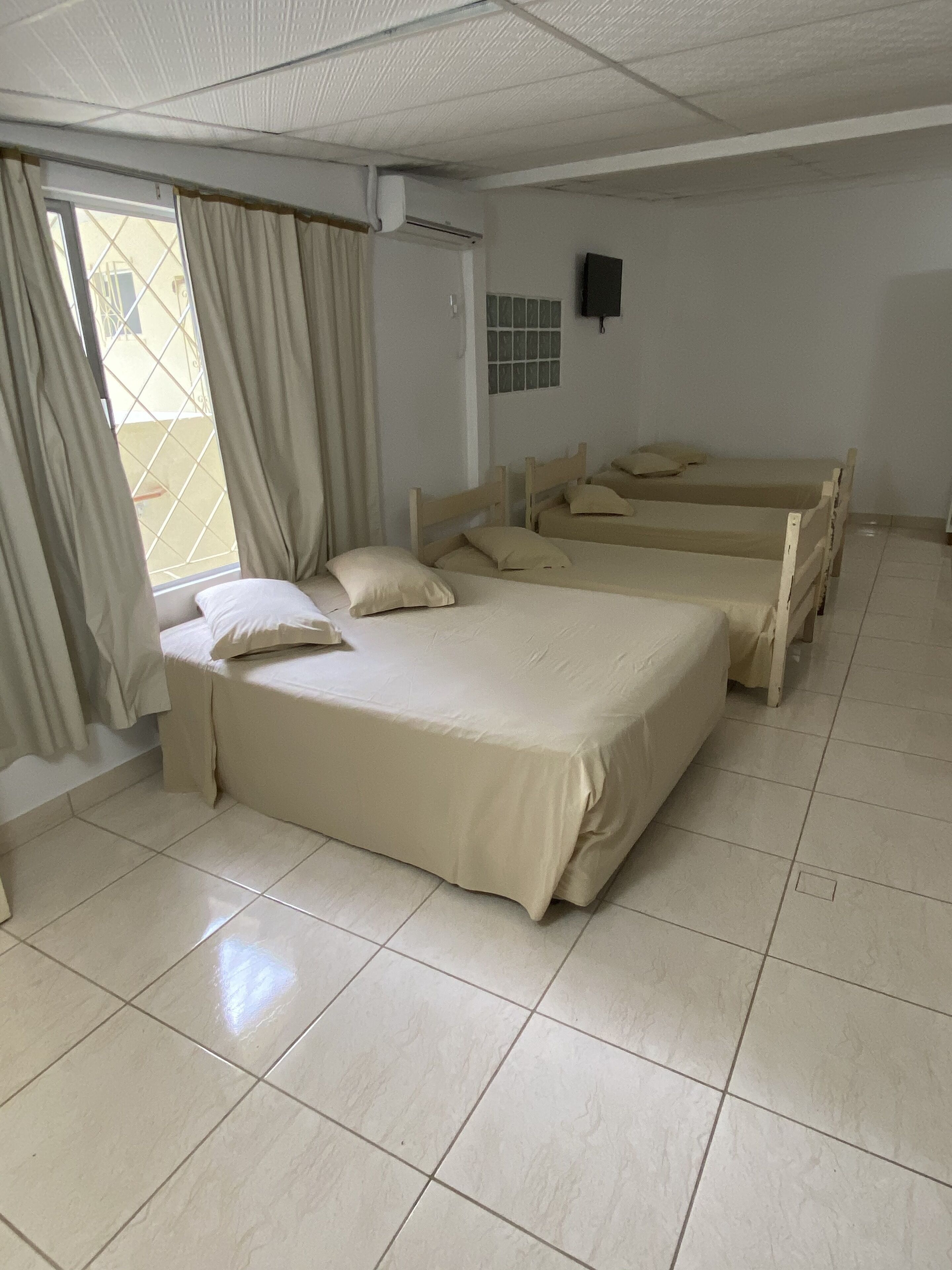 Quarto com três camas de madeira, chão de azulejos, janela com cortinas e ar condicionado fixado na parede.