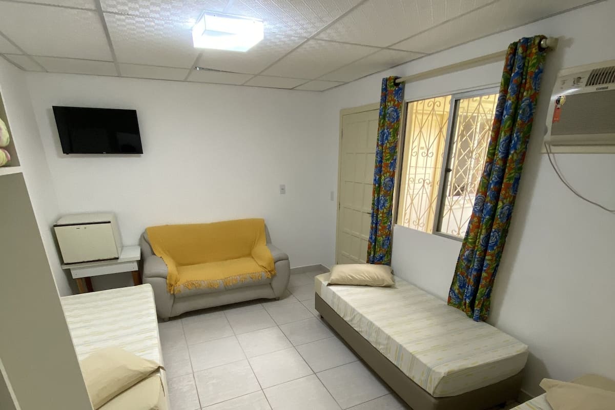 Habitación compacta con sofá, dos camas individuales, un escritorio pequeño con una computadora y un televisor en la pared.