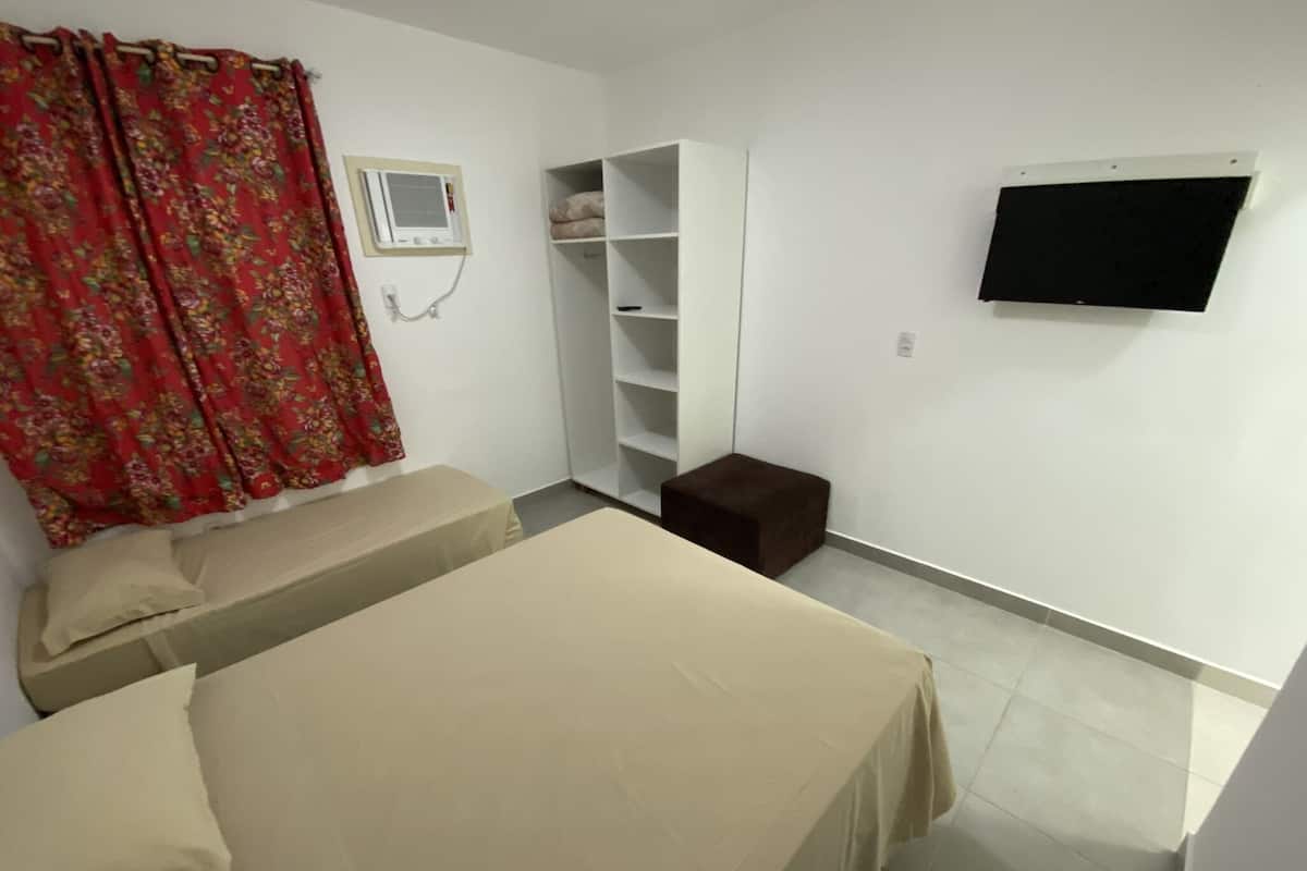 Una habitación con dos camas, un armario, un taburete pequeño, un televisor montado en la pared y una ventana con cortinas.