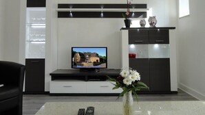 Una sala moderna con un televisor de pantalla plana, un mueble de entretenimiento oscuro, un jarrón blanco con flores y una vitrina de vidrio con copas de vino.