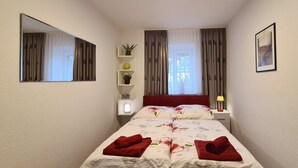 Una habitación de hotel con una cama, una mesita de noche, un espejo, una ventana con cortinas, un estante en la pared con plantas y un cuadro enmarcado.
