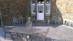Un patio con paredes de piedra, una mesa de vidrio y sillas de metal, una pequeña entrada con puerta blanca y plantas en macetas.