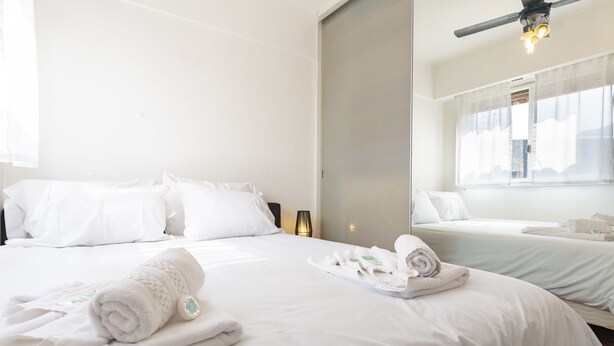 Une chambre d’hôtel avec deux lits, du linge de lit blanc, un ventilateur de plafond et une fenêtre avec des rideaux légers.