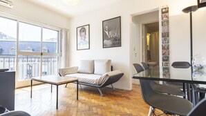 Un salon moderne avec un canapé, une table à manger en verre et une chaise. Deux tableaux encadrés ornent le mur, et une fenêtre offre une vue sur les bâtiments.