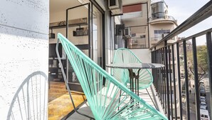 Un balcon avec une chaise verte et une petite table, donnant sur une rue bordée de voitures garées et d’immeubles.