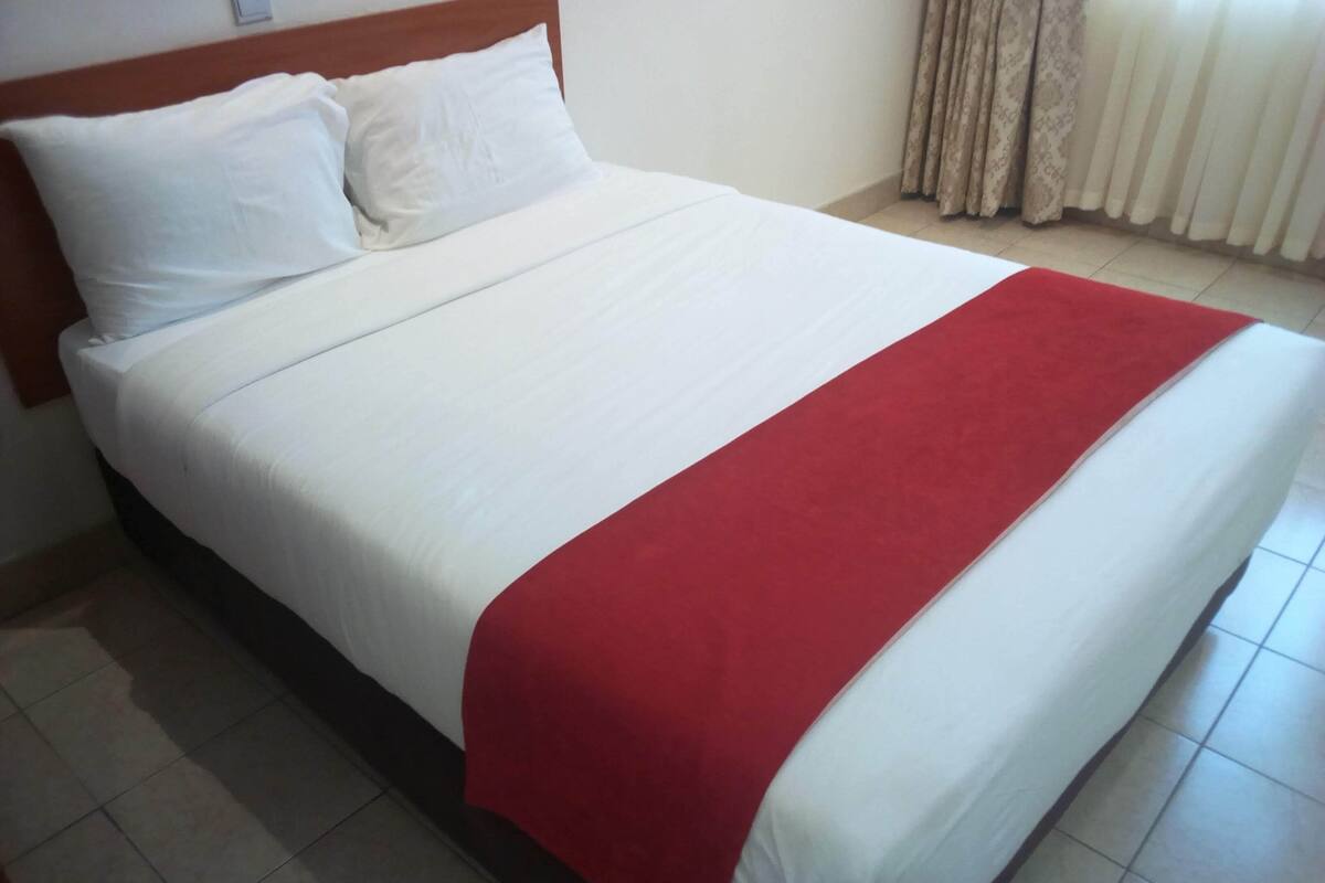 A neatly made bed with white bedding and a red blanket, a wooden headboard, and a tiled floor.