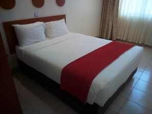 Una cama con ropa de cama blanca y un plaid rojo, un cabecero de madera y una pared lisa con un interruptor de luz.