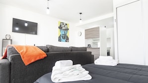 Una habitación de hotel moderna con un sofá gris, una cama con ropa blanca, un televisor de pantalla plana montado en la pared y una cocina visible a través de una puerta abierta.