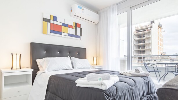 Una habitación de hotel moderna con una cama grande, mesitas de noche blancas, aire acondicionado empotrado en el techo y un balcón con vistas a un edificio en construcción.