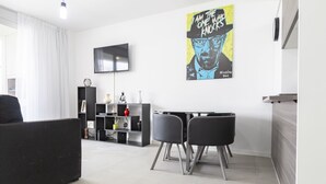 Una sala de estar moderna con un sofá negro, una mesa de comedor con dos sillas, un televisor montado en la pared y un cartel de un personaje de dibujos animados.