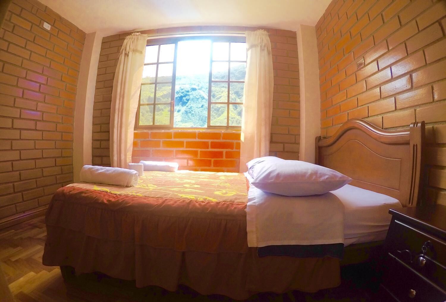 A room with a brick wall, a large window with curtains, a bed with pillows, and a wooden headboard.