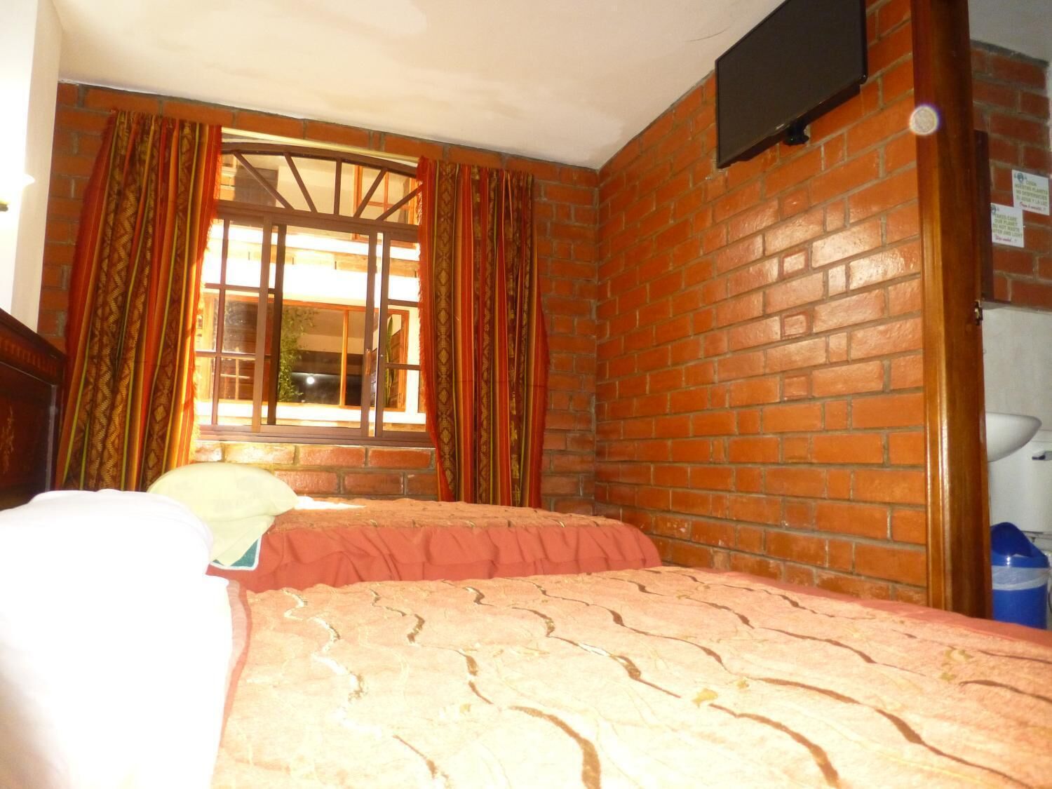 Una habitación con pared de ladrillo, ventana con cortinas, cama con colcha estampada y televisor montado en la pared.
