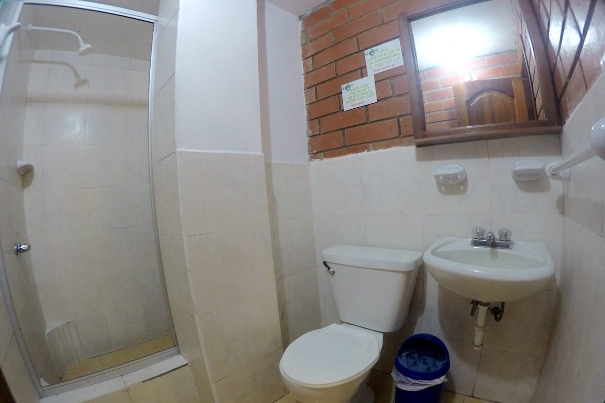 Une petite salle de bain avec des toilettes, un lavabo et une douche. Une poubelle bleue se trouve à côté du lavabo.