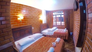 A room with two beds, each with a headboard, a window with curtains, and a brick wall. - Hostal Nomada - Hostel (Baños de Agua Santa)