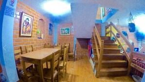 A dining area with a wooden table and chairs, a brick wall with artwork, and a staircase leading to an upper level. - Hostal Nomada - Hostel (Baños de Agua Santa)