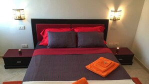 A neatly made bed with a red and grey striped bedspread, two pillows, and a folded orange towel. There are two bedside tables with lamps, and the room has a modern design. - Boa Vista Holidays (Boa Vista)