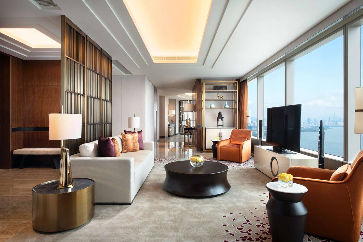 A modern hotel room with a sofa, armchairs, a coffee table, a TV, and a view of the city.