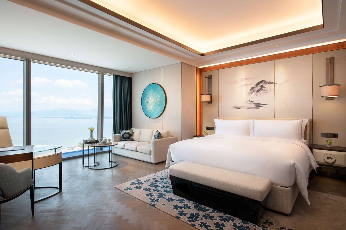 A modern hotel room with a large bed, a seating area with a sofa and armchair, a dining table, and a view of the sea.