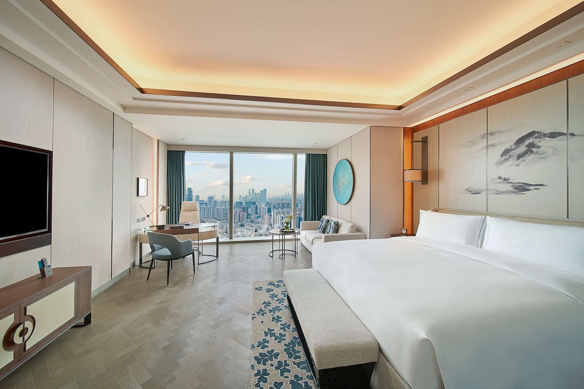 A modern hotel room with a large bed, a desk, a chair, and a television. The room has a city view through large windows.