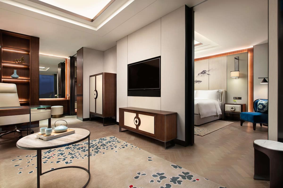 A modern hotel room with a flat-screen TV, a bed, a desk with a chair, a small table with a teapot, and a decorative rug.