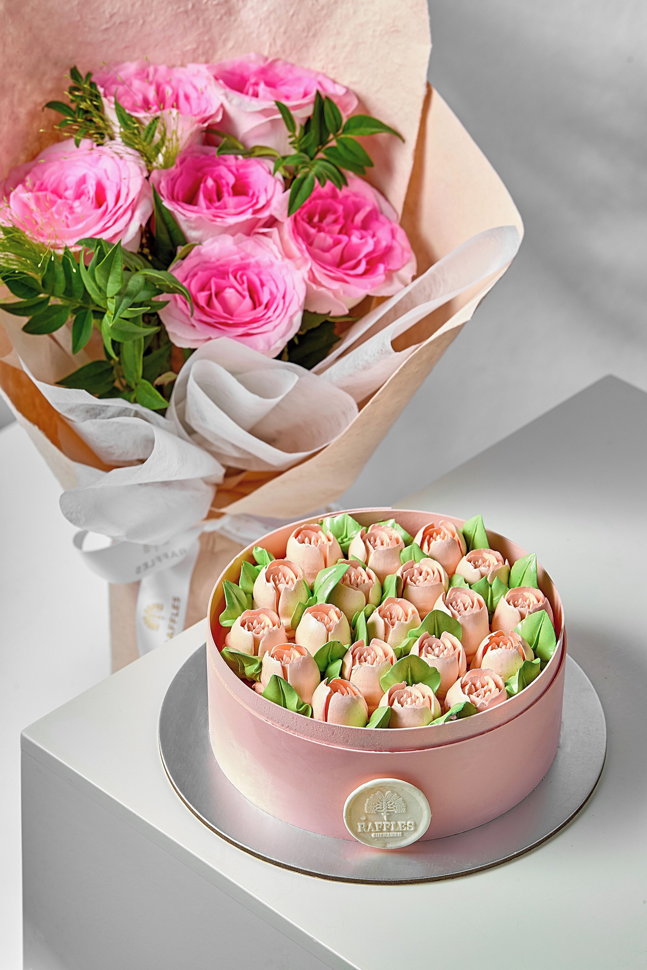 Um bolo cor-de-rosa com uma etiqueta sobre uma base prateada, acompanhado por um buquê de rosas cor-de-rosa em papel de embrulho.