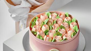 Um bolo cor-de-rosa com uma etiqueta sobre uma base prateada, acompanhado por um buquê de rosas cor-de-rosa em papel de embrulho.