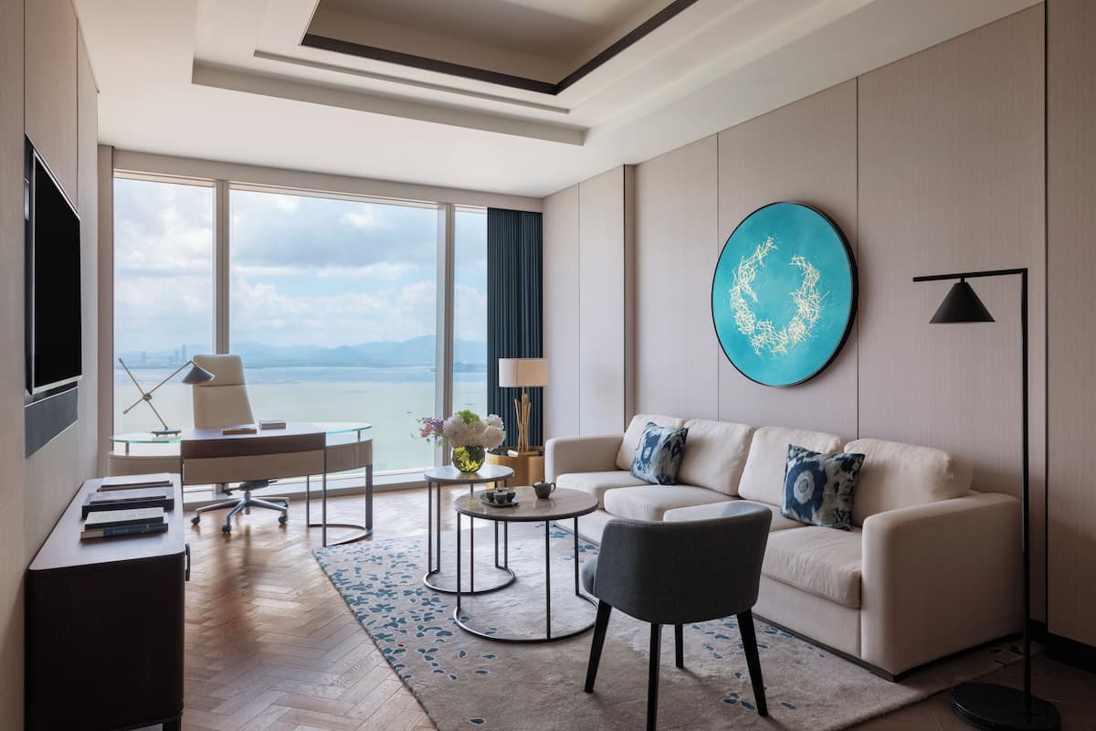 A modern living room with a sofa, coffee table, and TV, large windows with a view, and a decorative wall piece.
