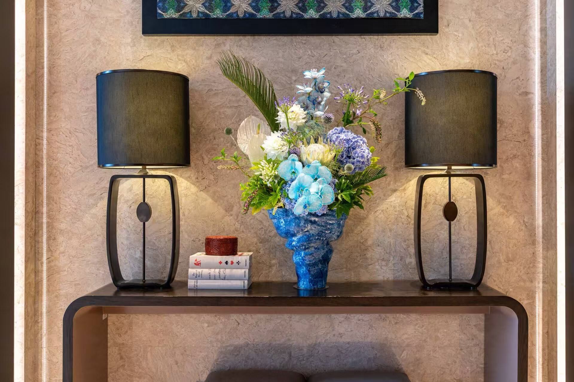 A room with a marble wall, a blue vase with flowers, two black lamps, and a small stack of books.