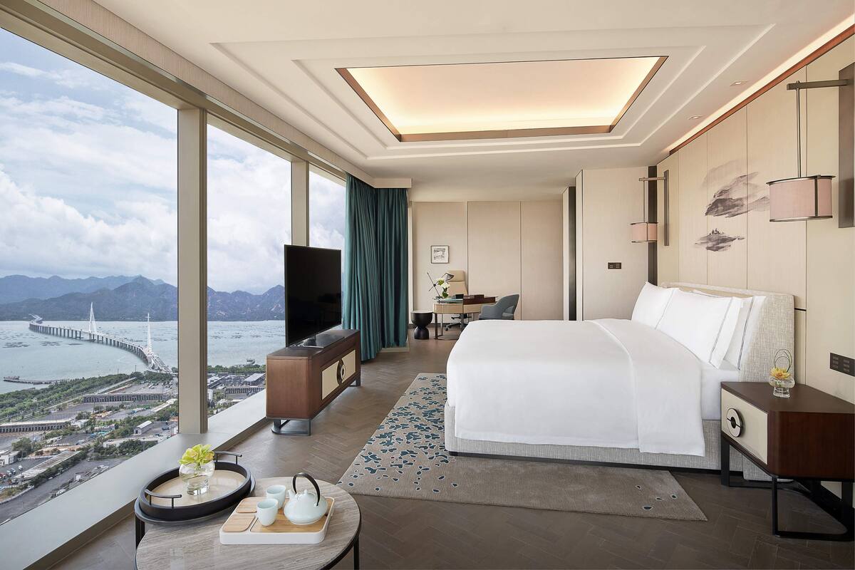 A hotel room with a large bed, a desk, a TV, and a view of a bridge and mountains.