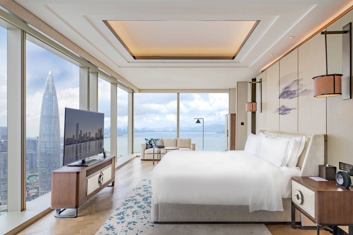 A modern hotel room with a large bed, a TV, a desk, and a view of a city skyline.