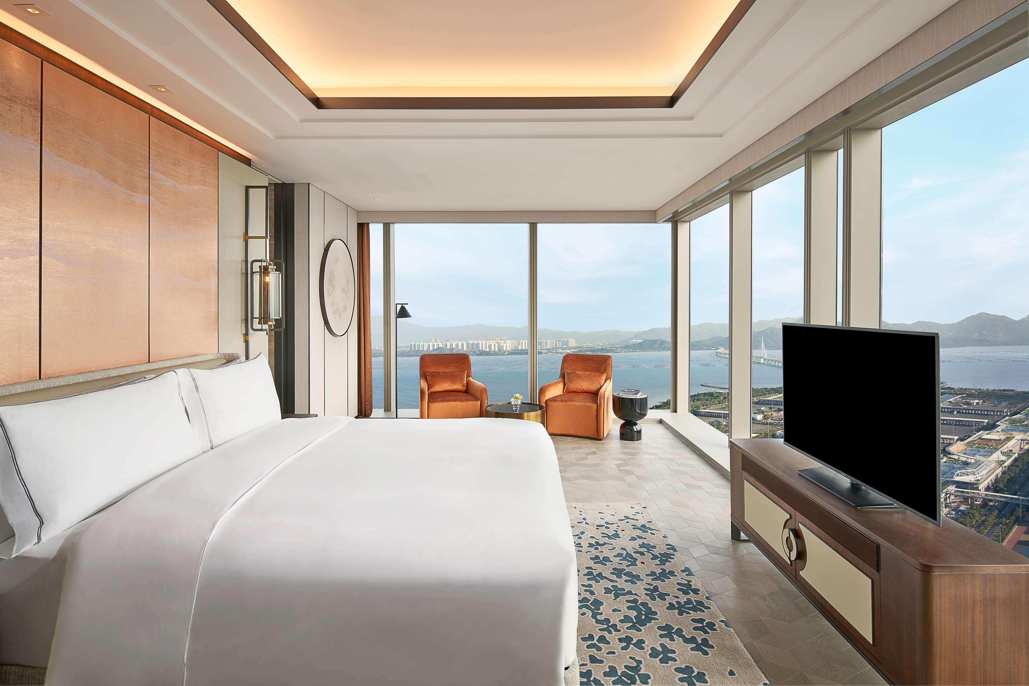 A modern hotel room with a large bed, two armchairs, a TV, and a view of a cityscape and water.