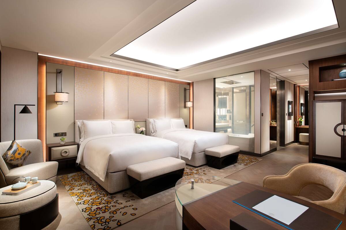 A hotel room with two beds, a desk, a chair, and a bathroom visible through a glass door.
