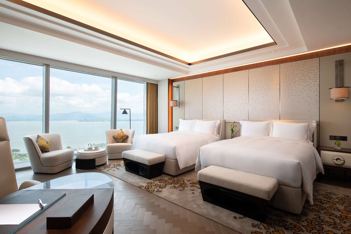 A hotel room with two beds, a desk, a chair, and a view of the sea.