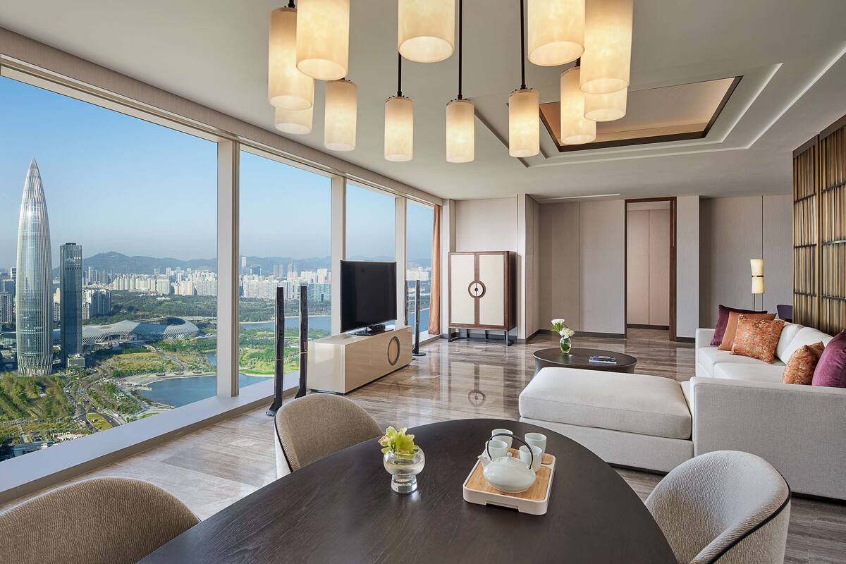 A modern living room with a large window offering a city view, a dining area with a table set for two, and a seating area with a sofa.