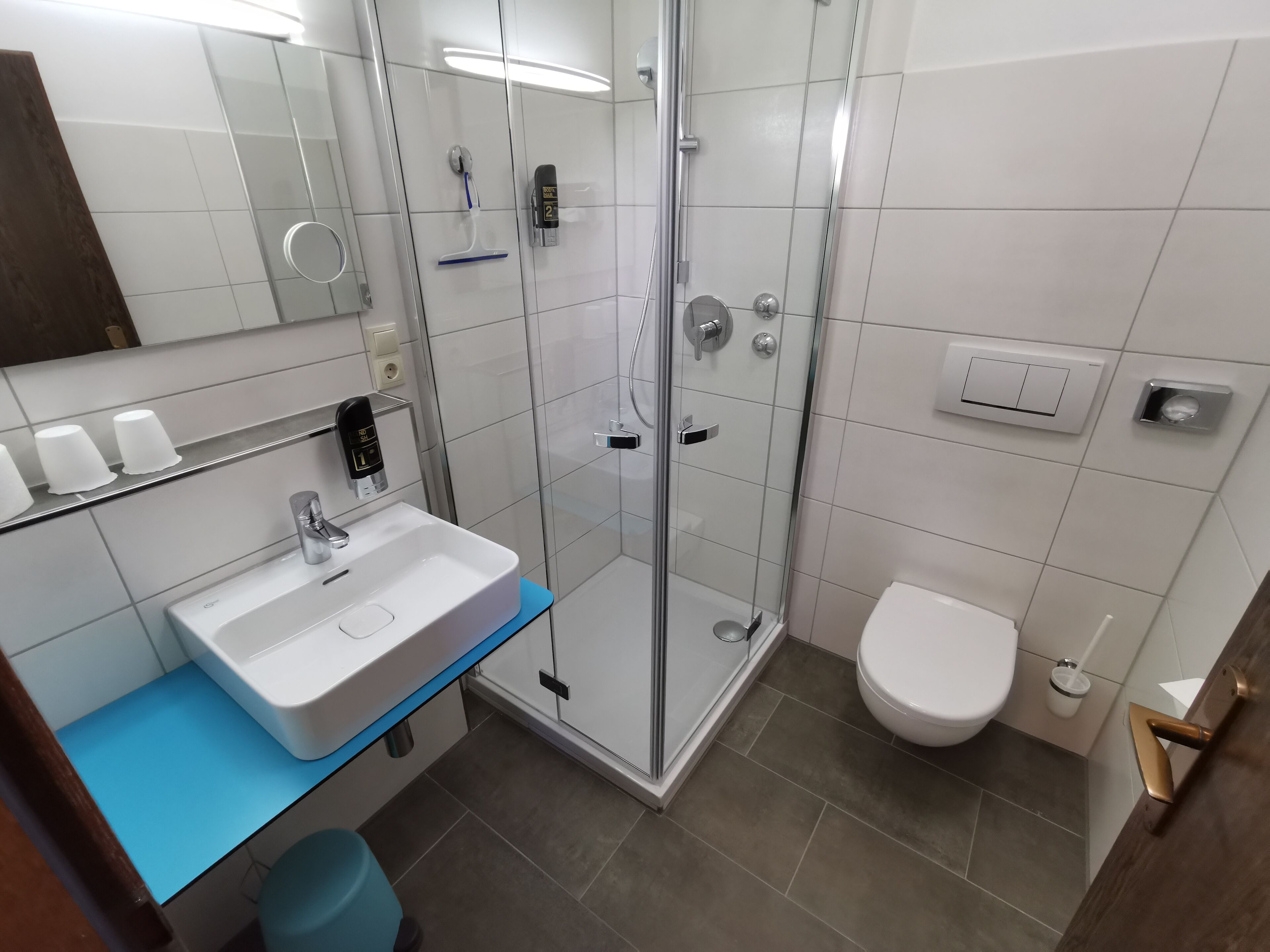 Een moderne badkamer met een glazen douchecabine, een witte wastafel met een blauw aanrechtblad, een toilet en een spiegel boven de wastafel.