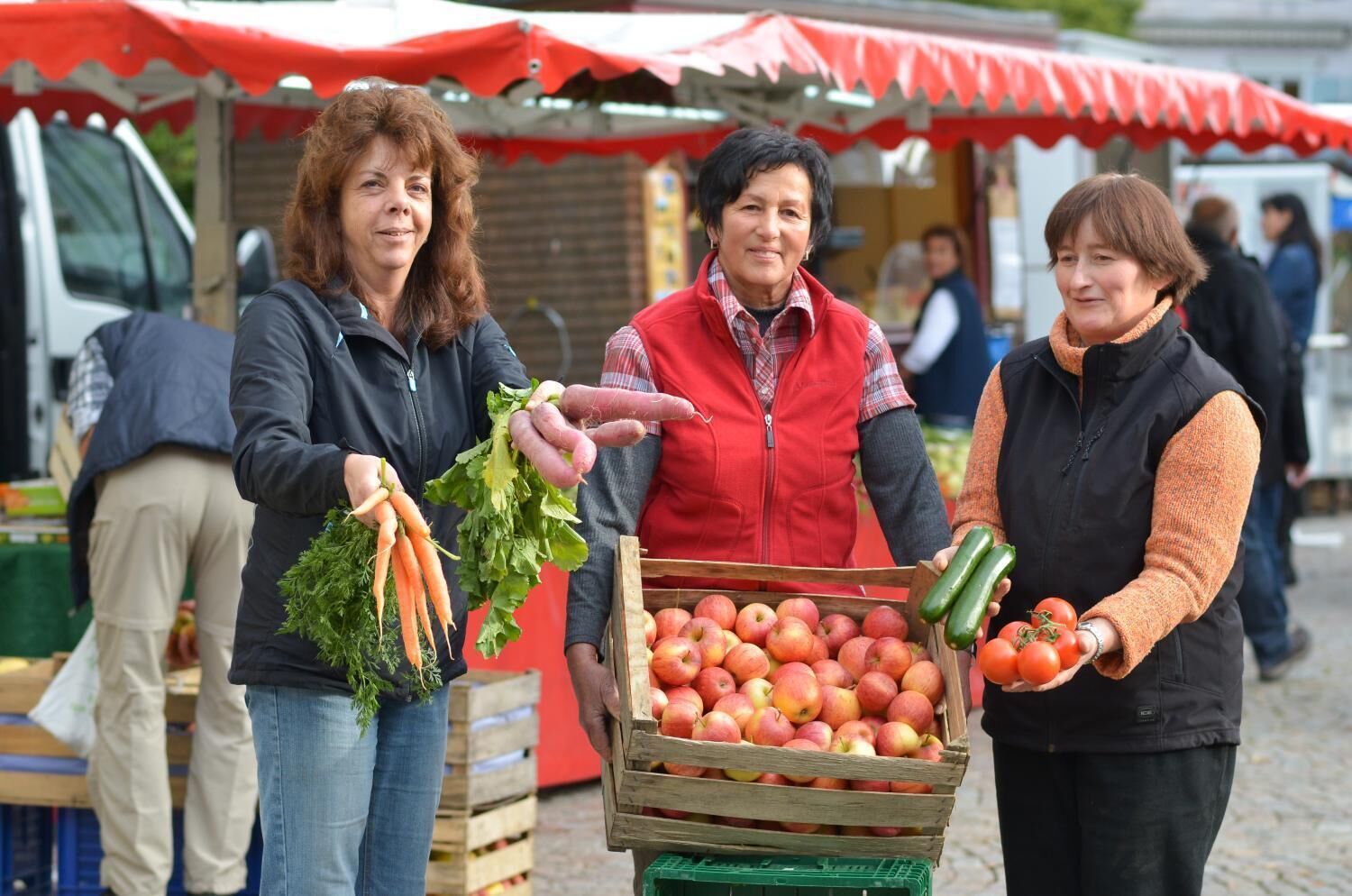 Drie personen op een openbare markt, met kisten vol appels en groenten.
