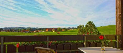 Een balkon met een tafel en stoelen, met uitzicht op een groen landschap met huizen en een heldere lucht.