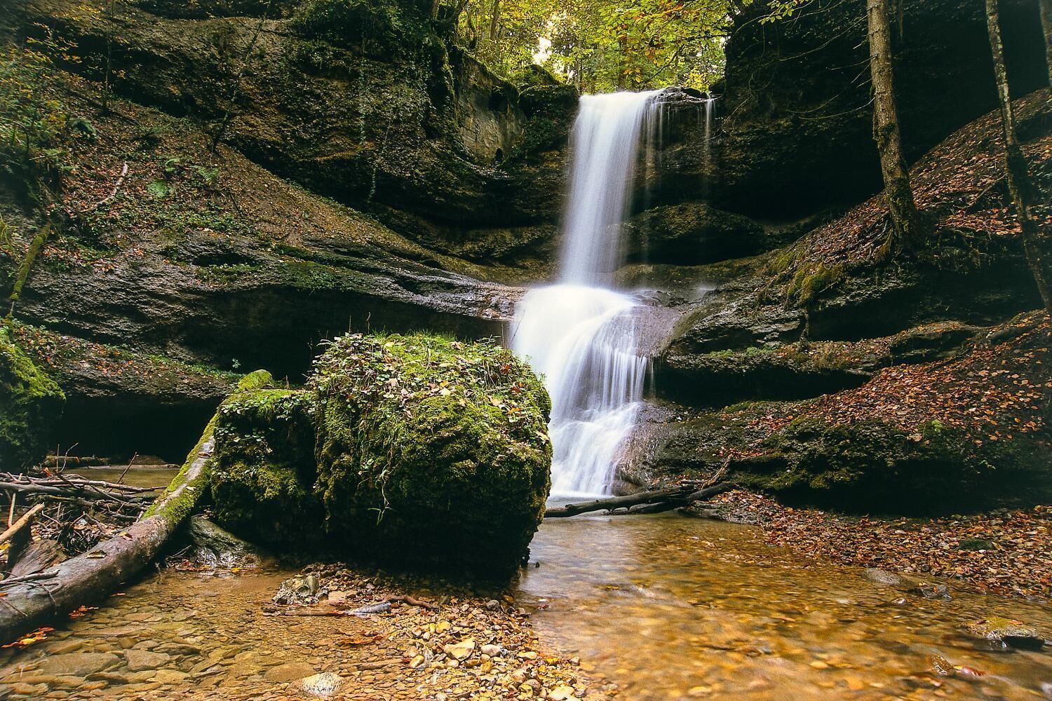 Une cascade située dans une zone boisée, avec des rochers couverts de mousse et un ruisseau aux eaux claires à son pied.