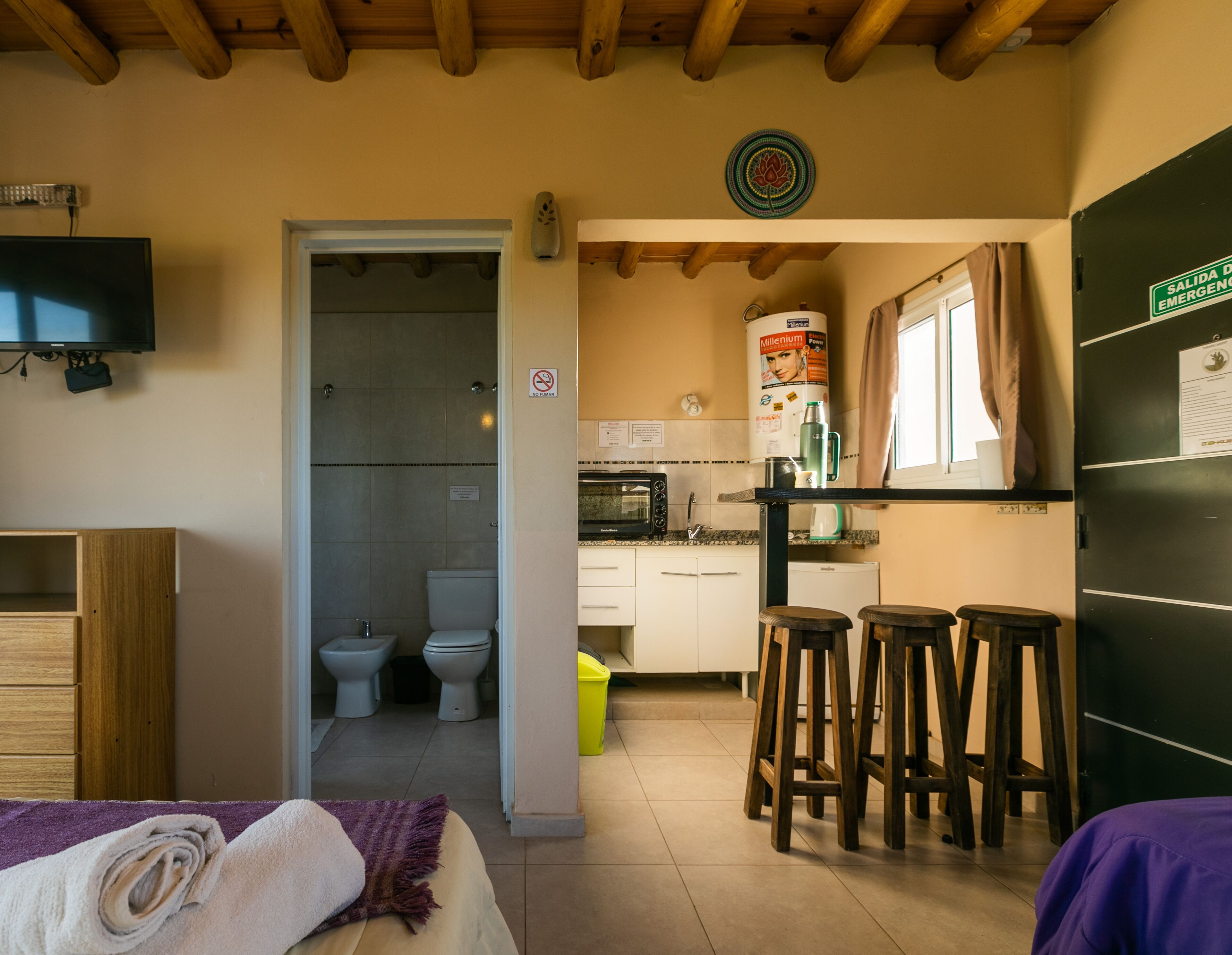 Camera d'albergo compatta con soffitto in legno, un letto con un asciugamano arrotolato, angolo cottura con forno a microonde, macchina del caffè e frigorifero. Nella zona cucina si trovano anche sgabelli di legno e un cestino per la spazzatura.