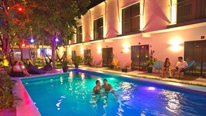 Zona de piscina con un edificio al fondo, rodeada de plantas en macetas y mobiliario exterior.